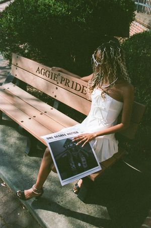 Someone in a white dress sits on an 'Aggie Pride' bench while holding what appears to be a photo album.
