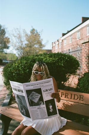A graduate sits outside reading a newspaper in a white dress with sunglasses and Pride decor visible in the background.