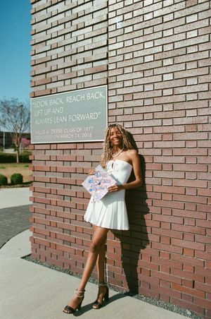 A person in a white dress poses against a brick wall next to a building signage plaque.