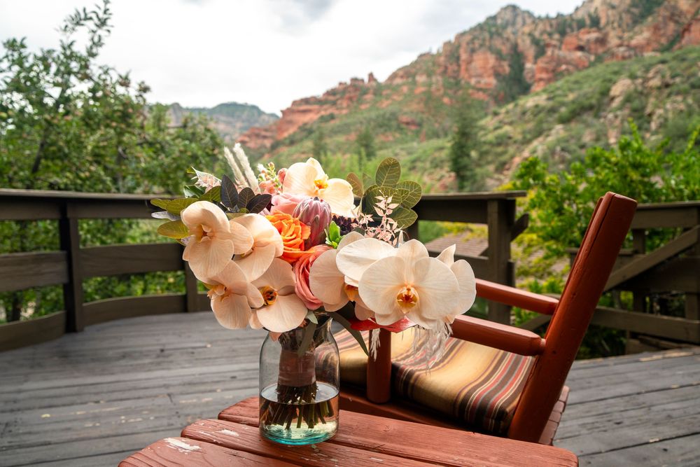 Floral arrangement with white orchids on a wooden deck overlooking red rock mountains and lush greenery.