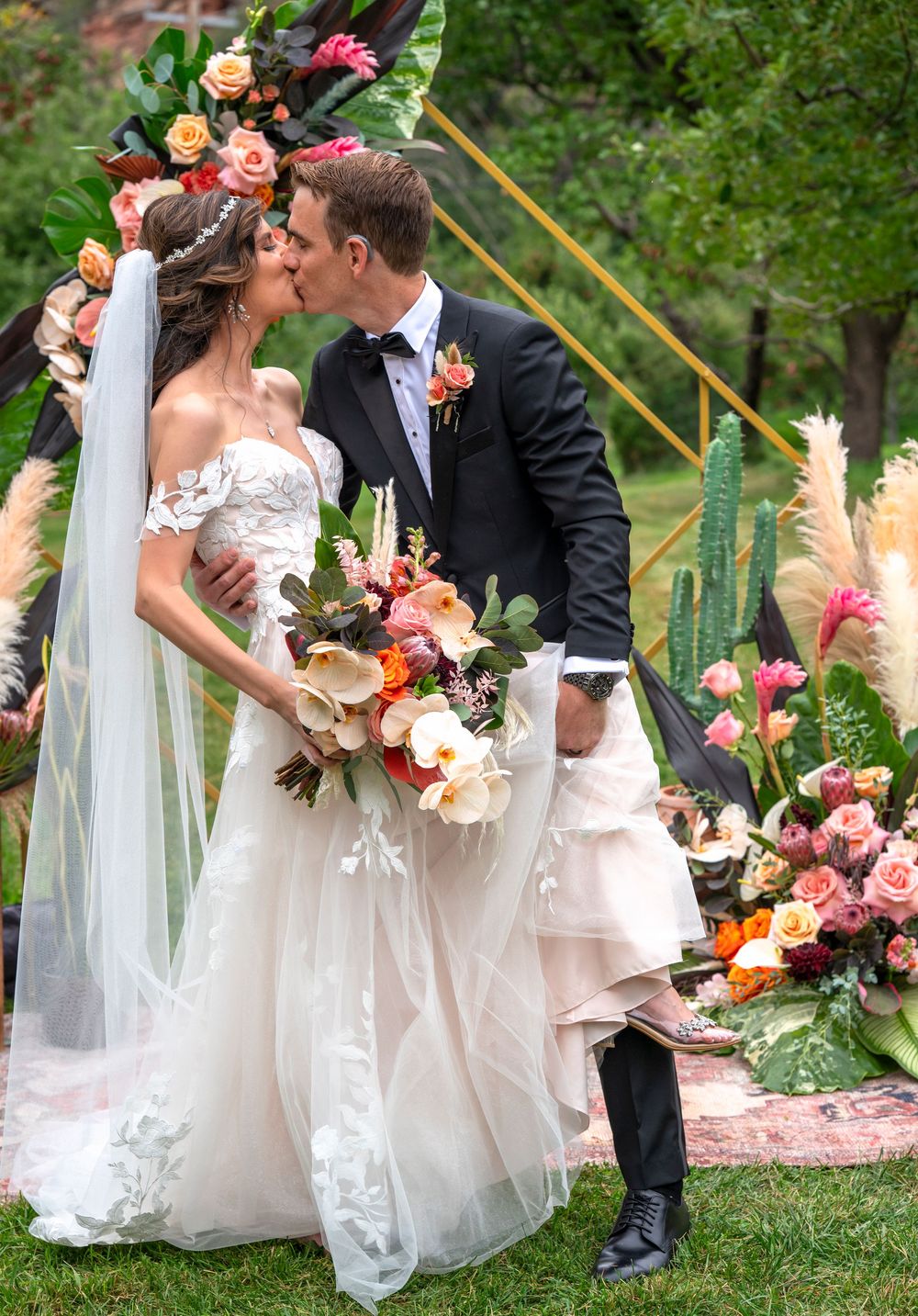 Bride and groom share a kiss at outdoor wedding ceremony with colorful floral arch and lush bouquet.