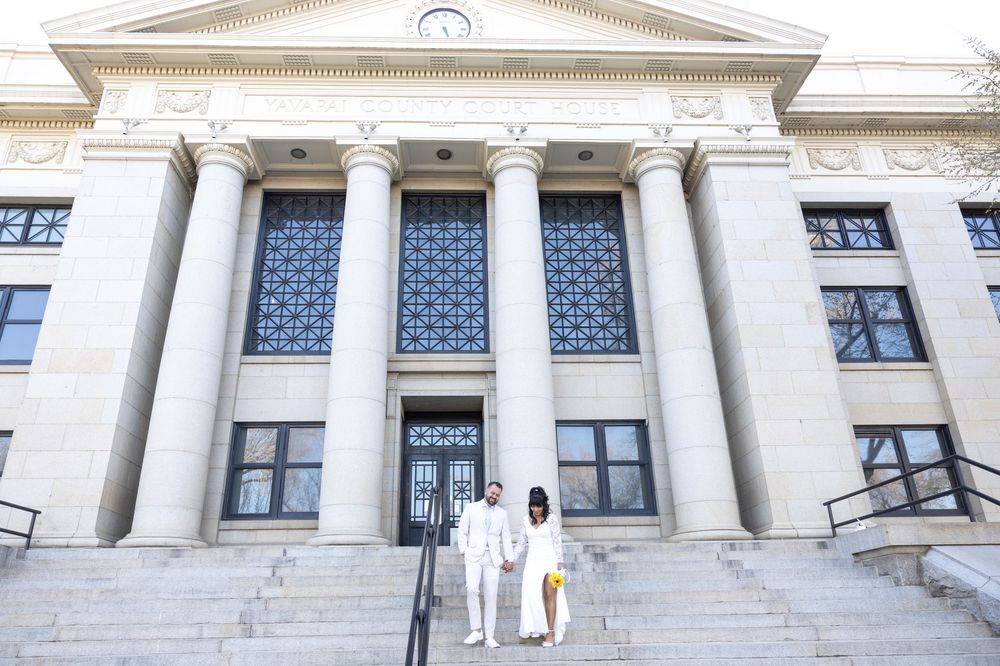 Two figures stand on the grand steps of a neoclassical building with white columns and triangular pediment.