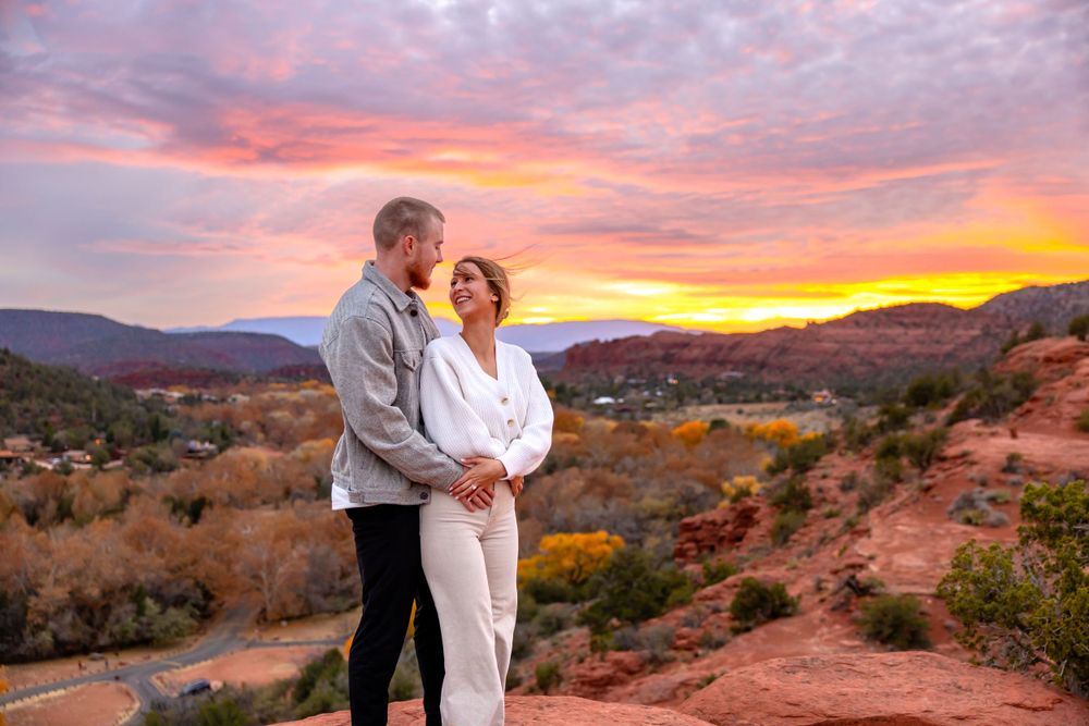 A couple embraces on red rocks overlooking a desert landscape during a vibrant sunset in the mountains. Sedona Engagement Photography