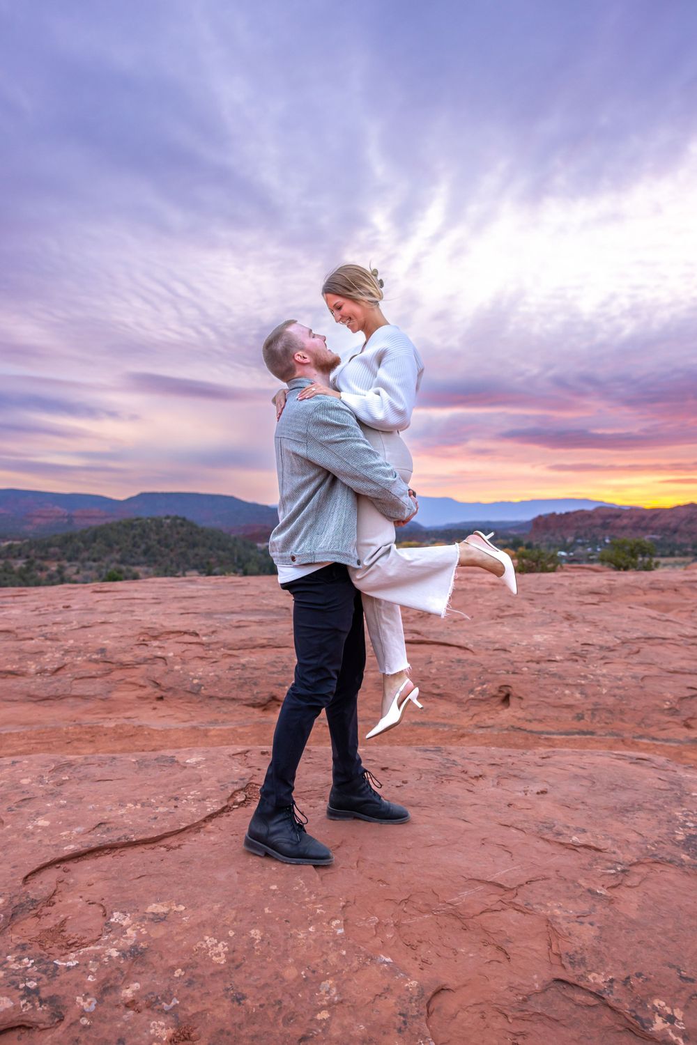 Two people embrace on red rocks at sunset with purple and orange clouds swirling in the desert sky. Sedona Engagement Photography