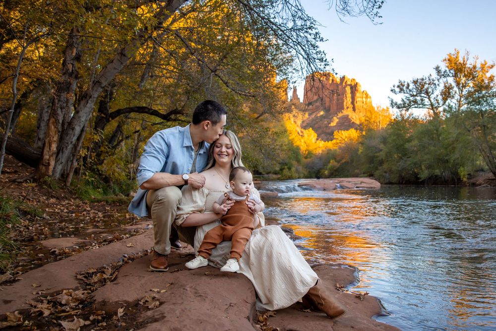 Family of three sitting on red rocks by a river in Sedona, Arizona, surrounded by autumn foliage and red rock formations.