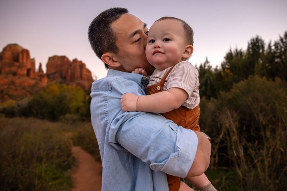 A father kisses his smiling baby outdoors near red rock formations at sunset in Sedona, Arizona.