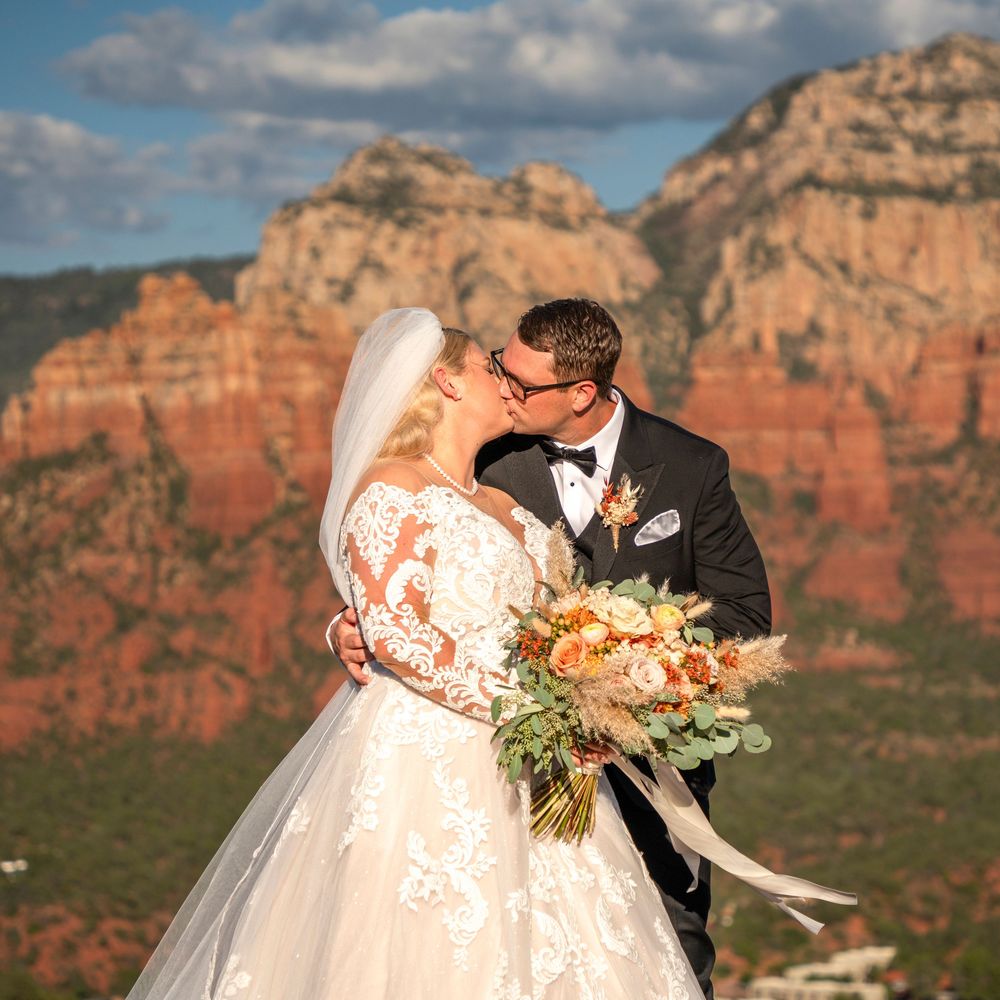 Bride and groom kissing with stunning Sedona red rock formations backdrop, bride in lace gown holding autumn bouquet.
