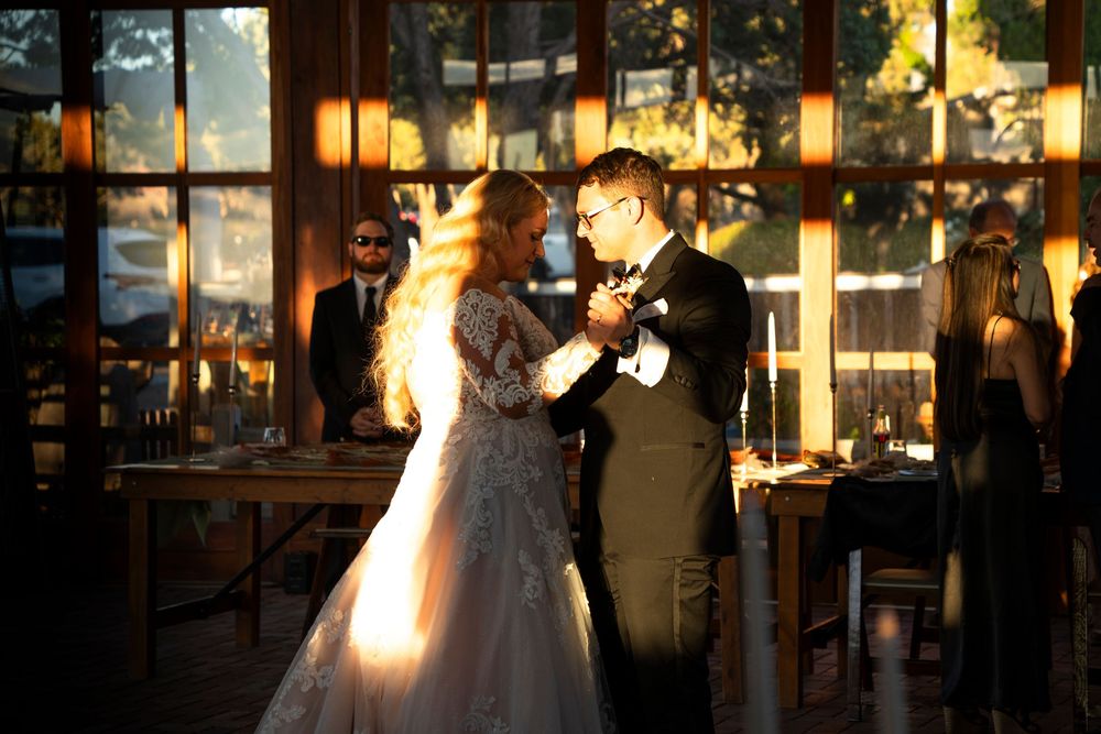 Bride and groom share their first dance in a warmly lit rustic wedding venue with large windows.