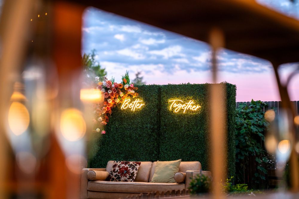 Romantic outdoor lounge with green hedge wall, neon "Better Together" sign, and floral accents at sunset.