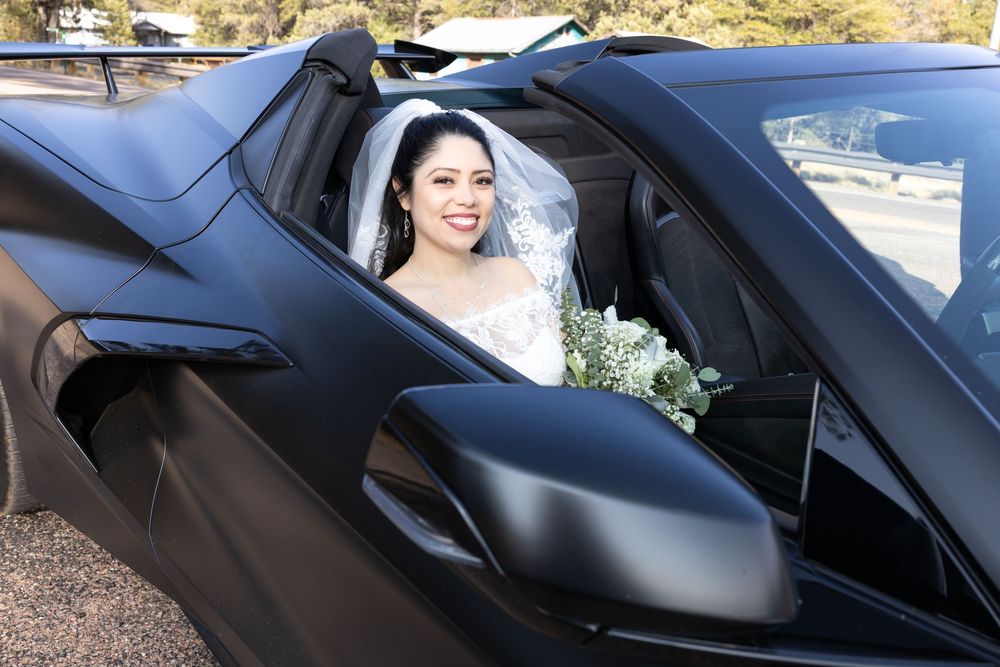 A bride in a white wedding dress and veil sits in a sleek black sports car holding a floral bouquet.