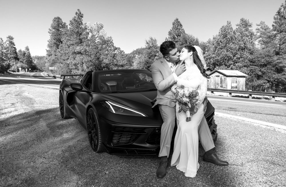 A couple shares a romantic moment leaning against a black Chevrolet Corvette C8 on a sunny day.