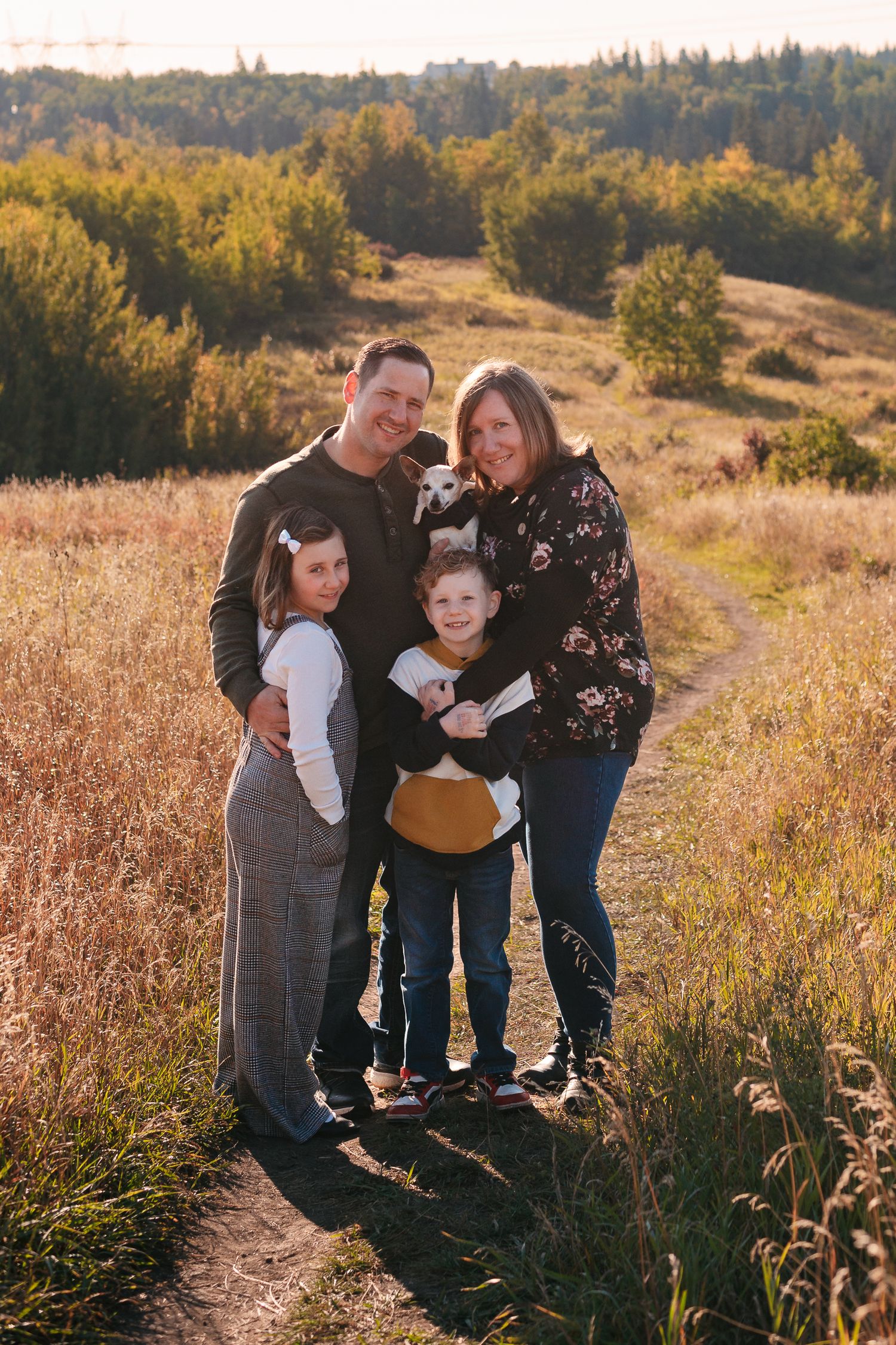 Family portrait in a golden field at sunset with children and a small white dog.