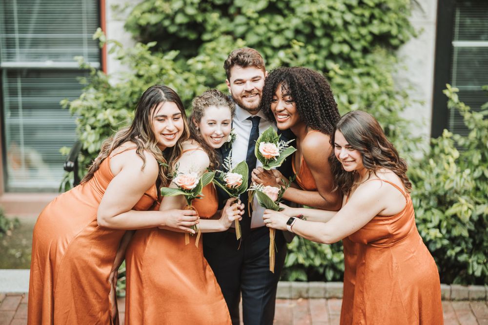 Bridal party in orange dresses sharing laughs while holding bouquets outdoors.
