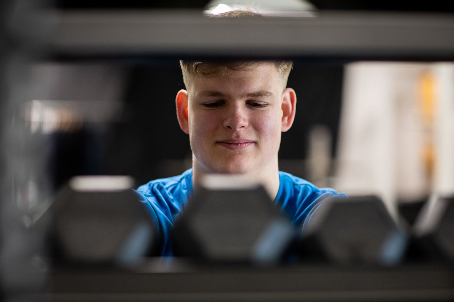 A person viewed through exercise equipment rack with dumbbells at a gym during business branding photoshoot.