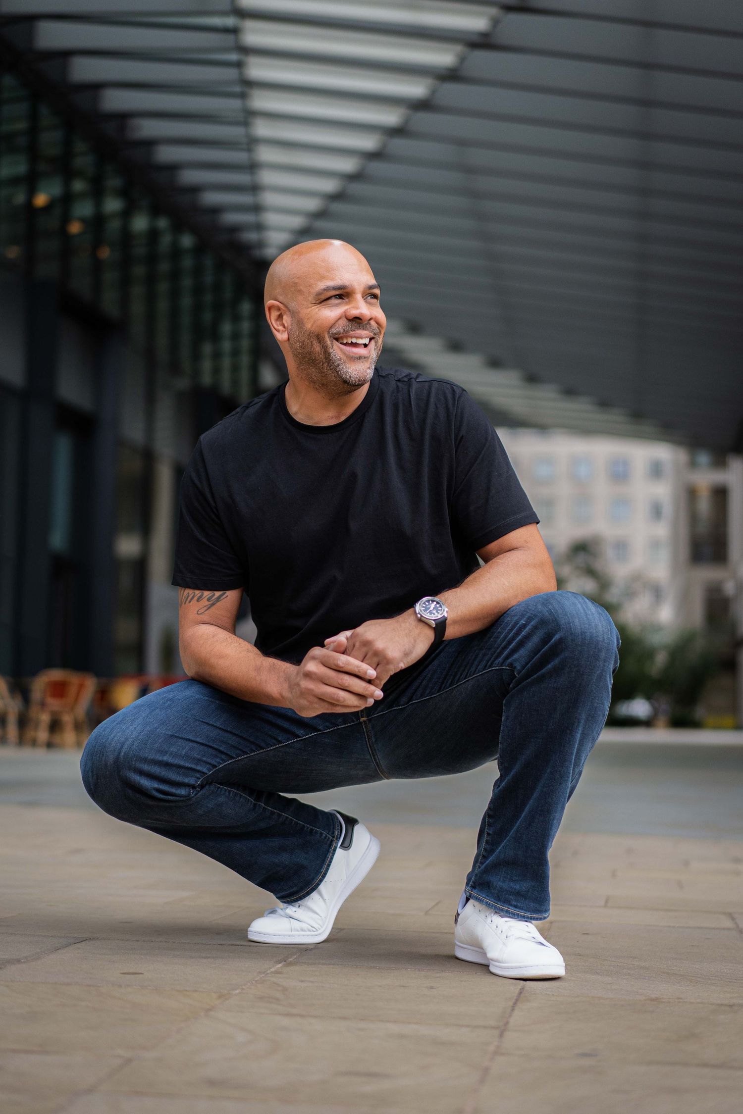 Man in black t-shirt and jeans crouches and smiles in modern architectural setting.