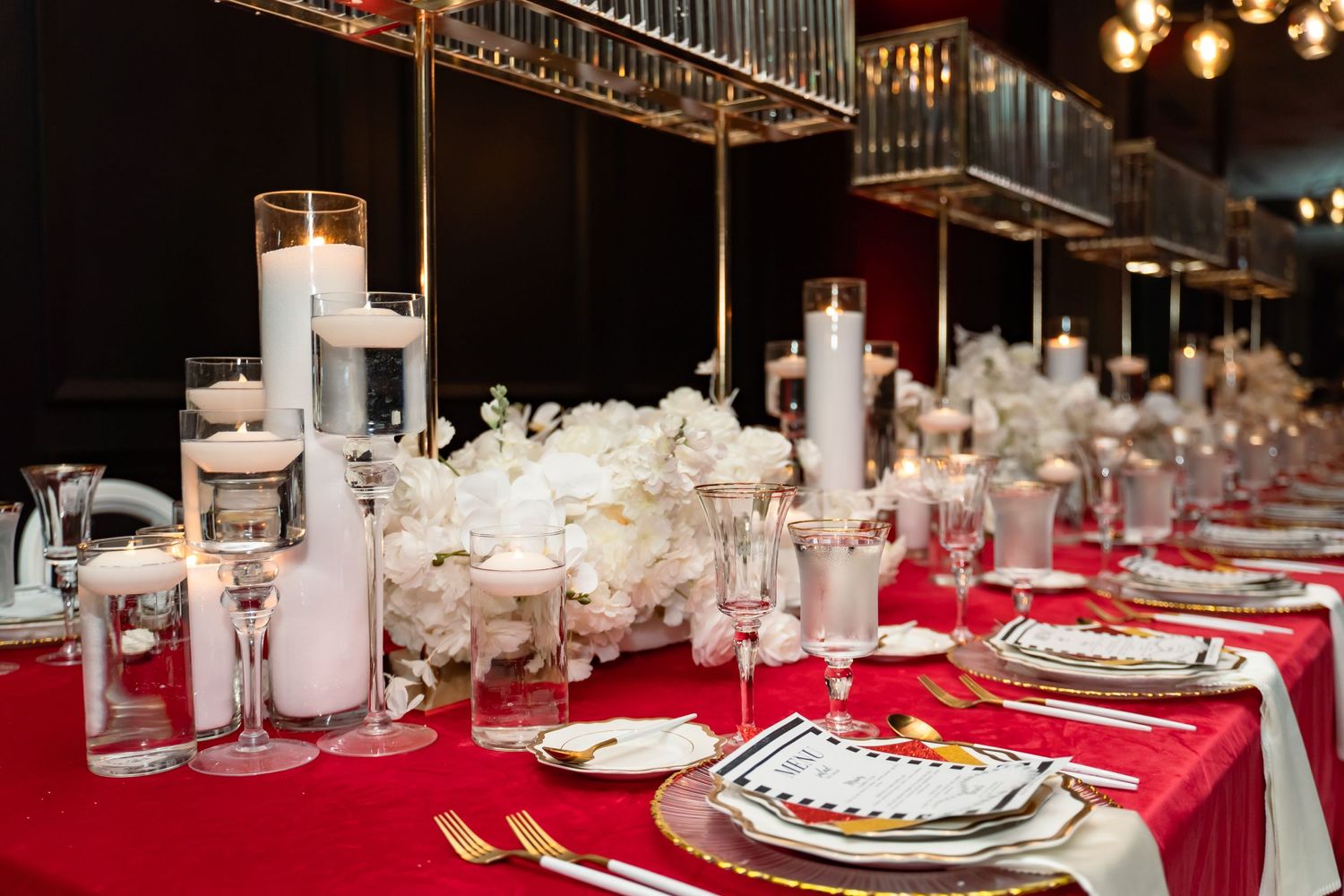 Elegant wedding reception table setting with white floral centerpieces, candles and red tablecloth.
