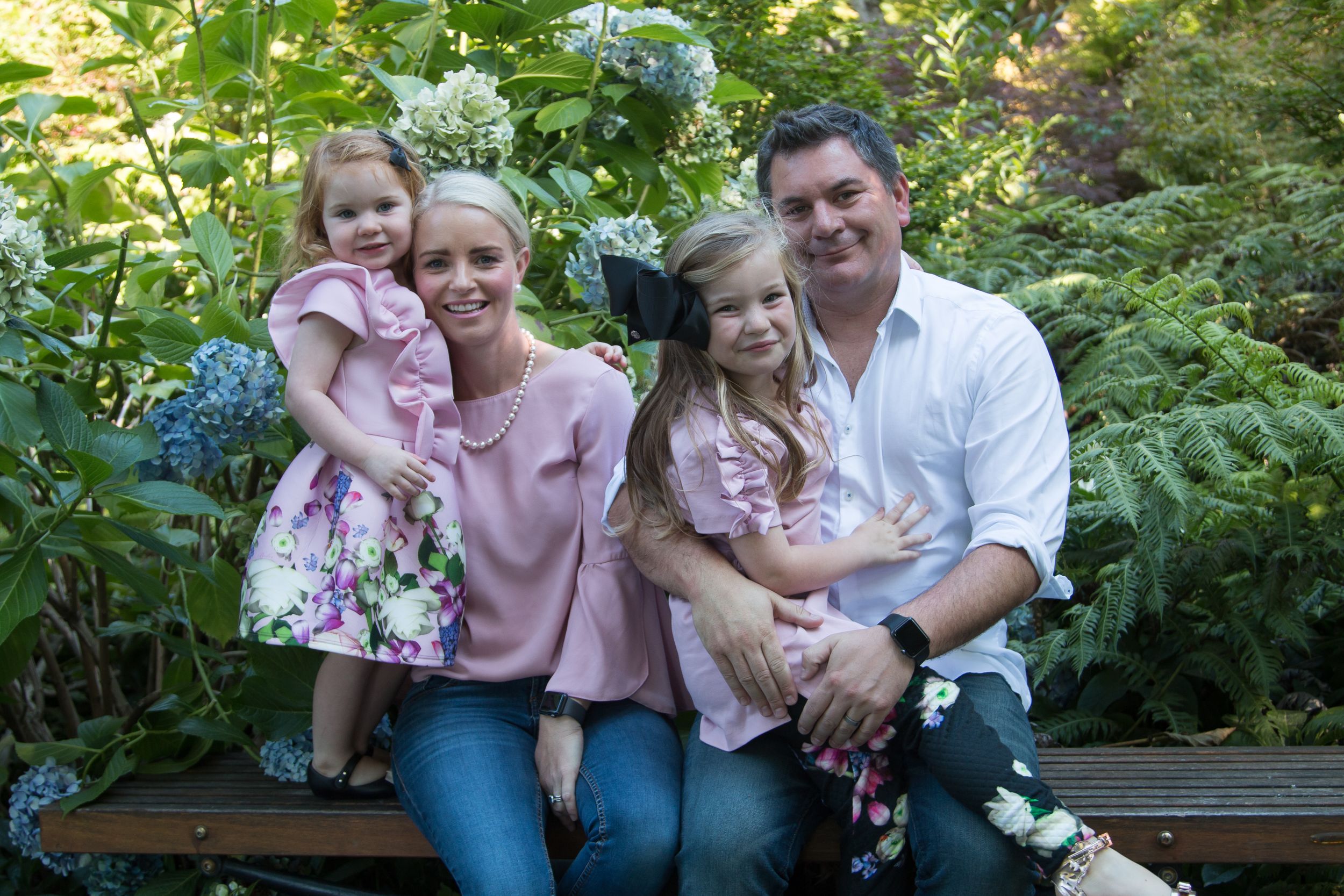 A family poses together on a wooden bench in a garden setting with blooming flowers.