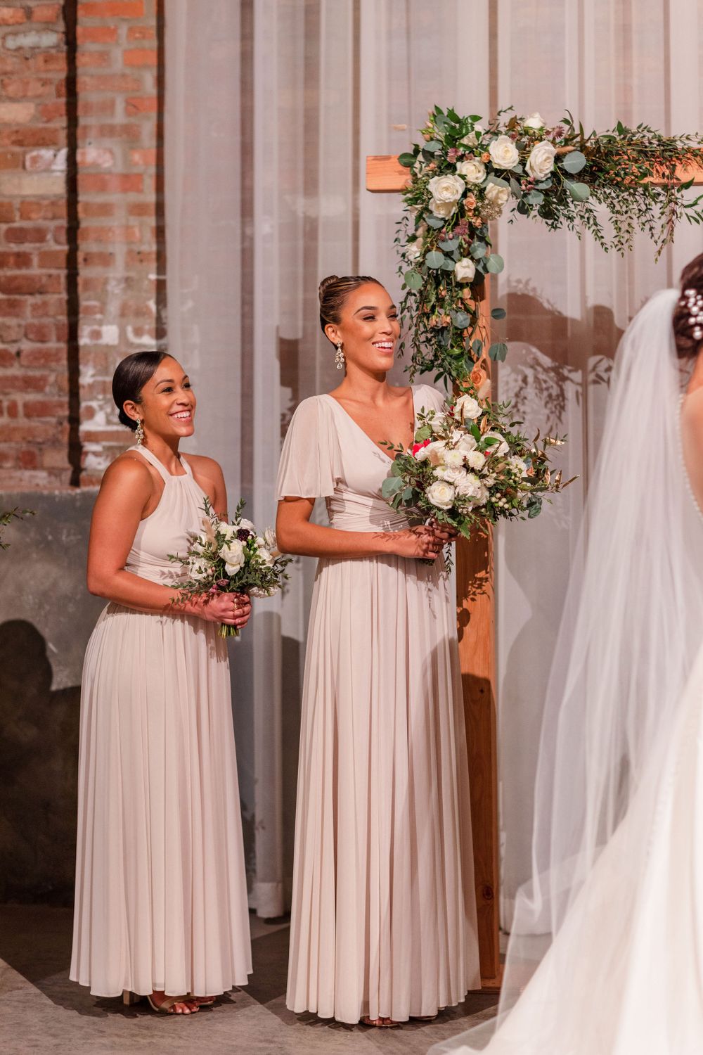 Bridesmaids in blush gowns holding white bouquets stand beside a draped floral arch.
