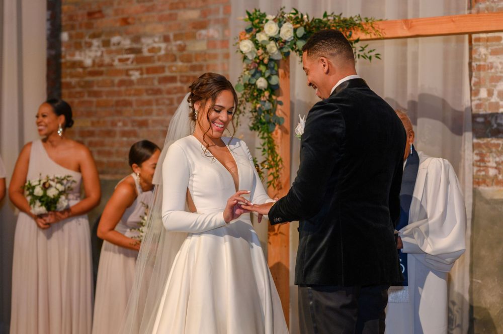 A wedding ceremony with a ring exchange in front of a floral arch against exposed brick walls.