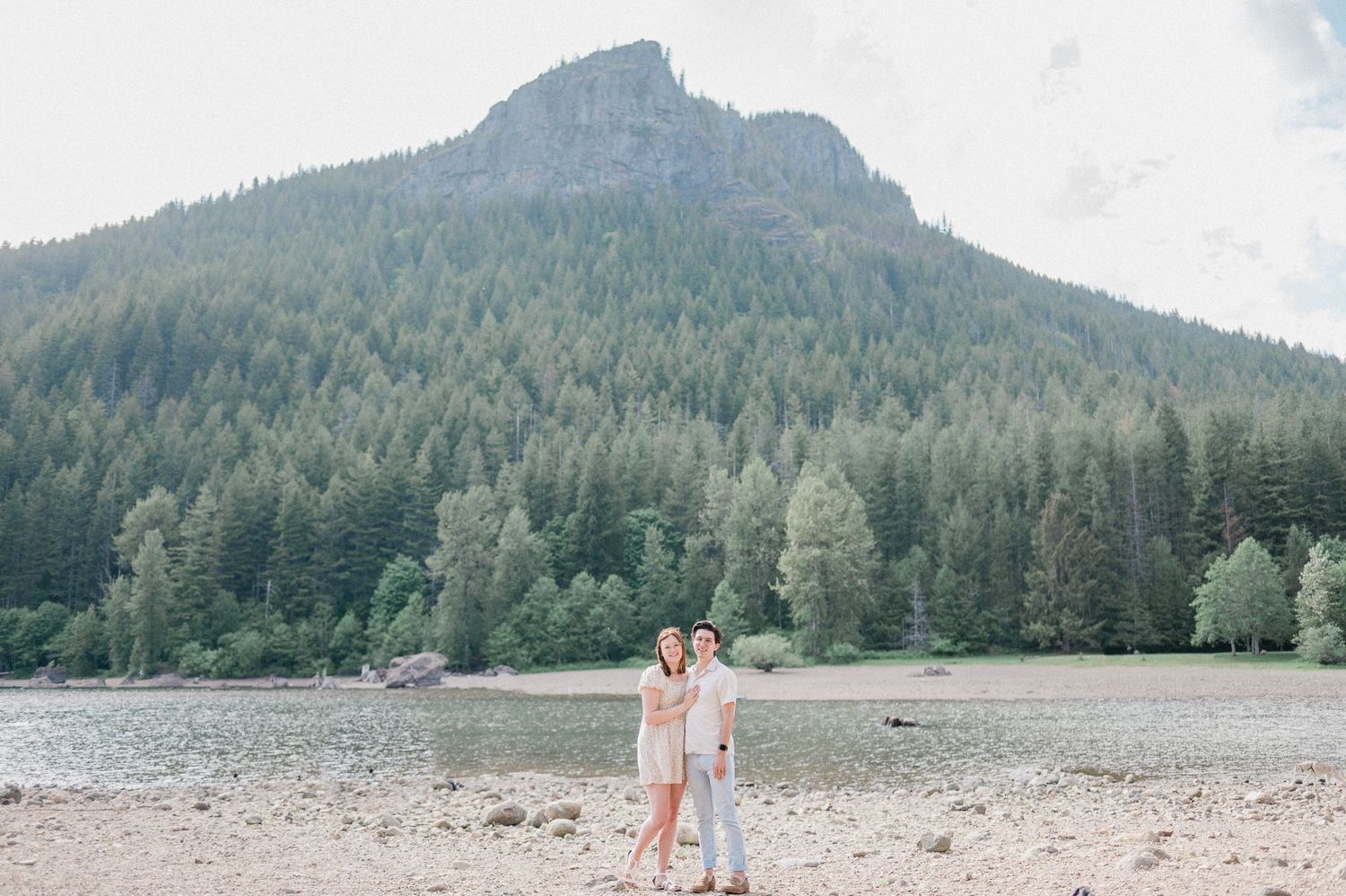 A couple stands together on a rocky beach with evergreen-covered mountains and cloudy skies in the background.