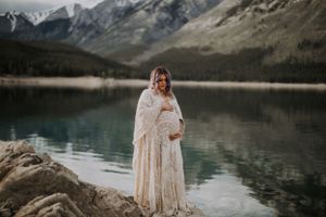 A woman in a lace dress stands by a serene mountain lake with snow-capped peaks in the background.