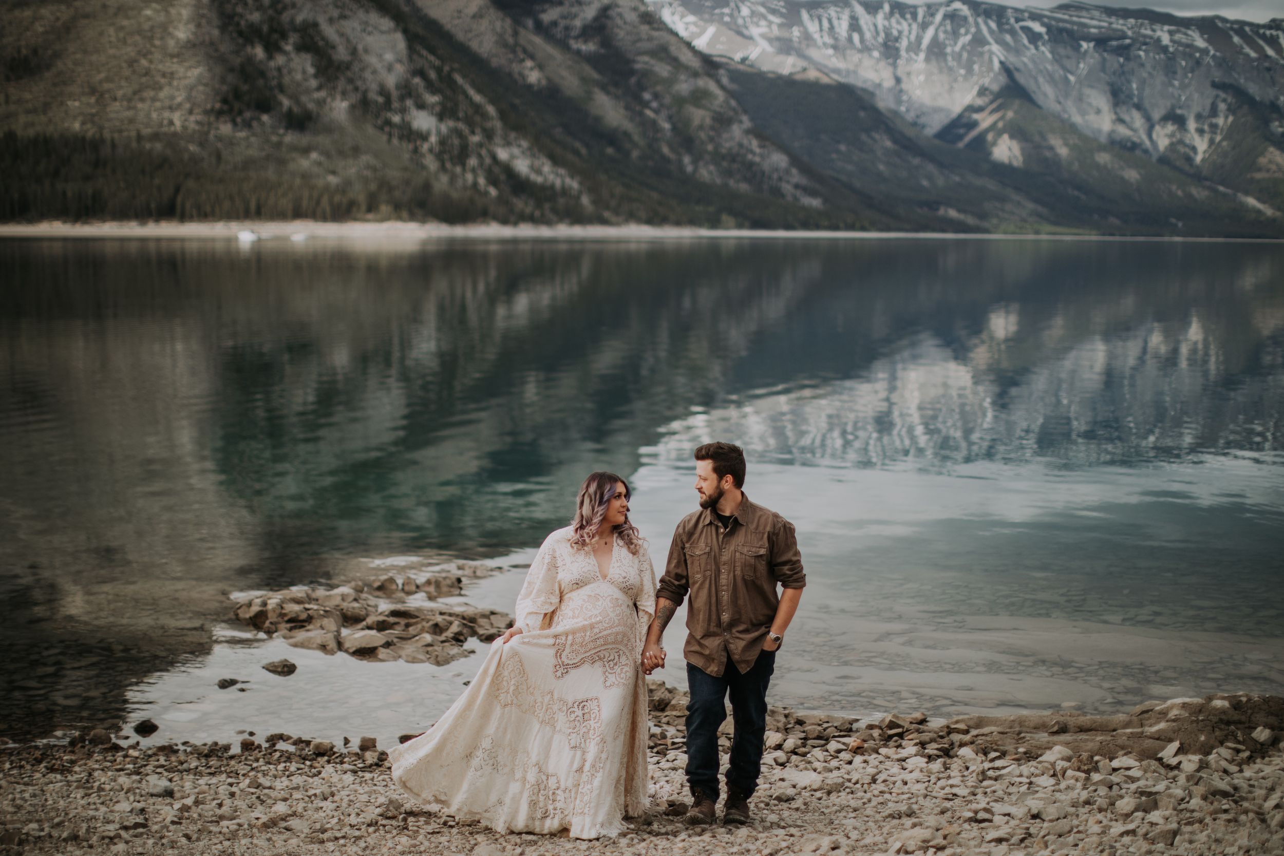 Couple stands together on rocky lakeshore with dramatic mountain landscape and reflective water.