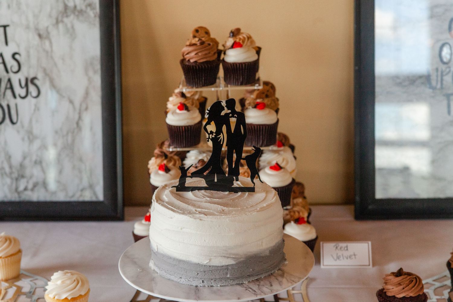 White wedding cake topped with chocolate frosted cupcakes and decorative toppers on a dessert table.