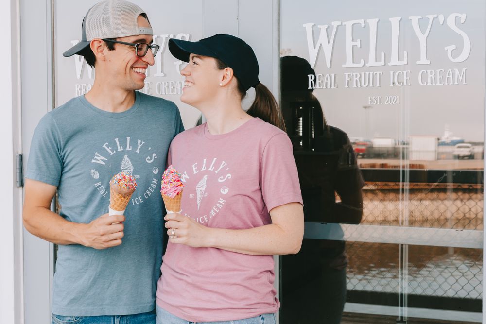 A couple look at each other while holding ice cream cones wearing shirts that say Welly’s Real Fruit Ice Cream infront of door says same