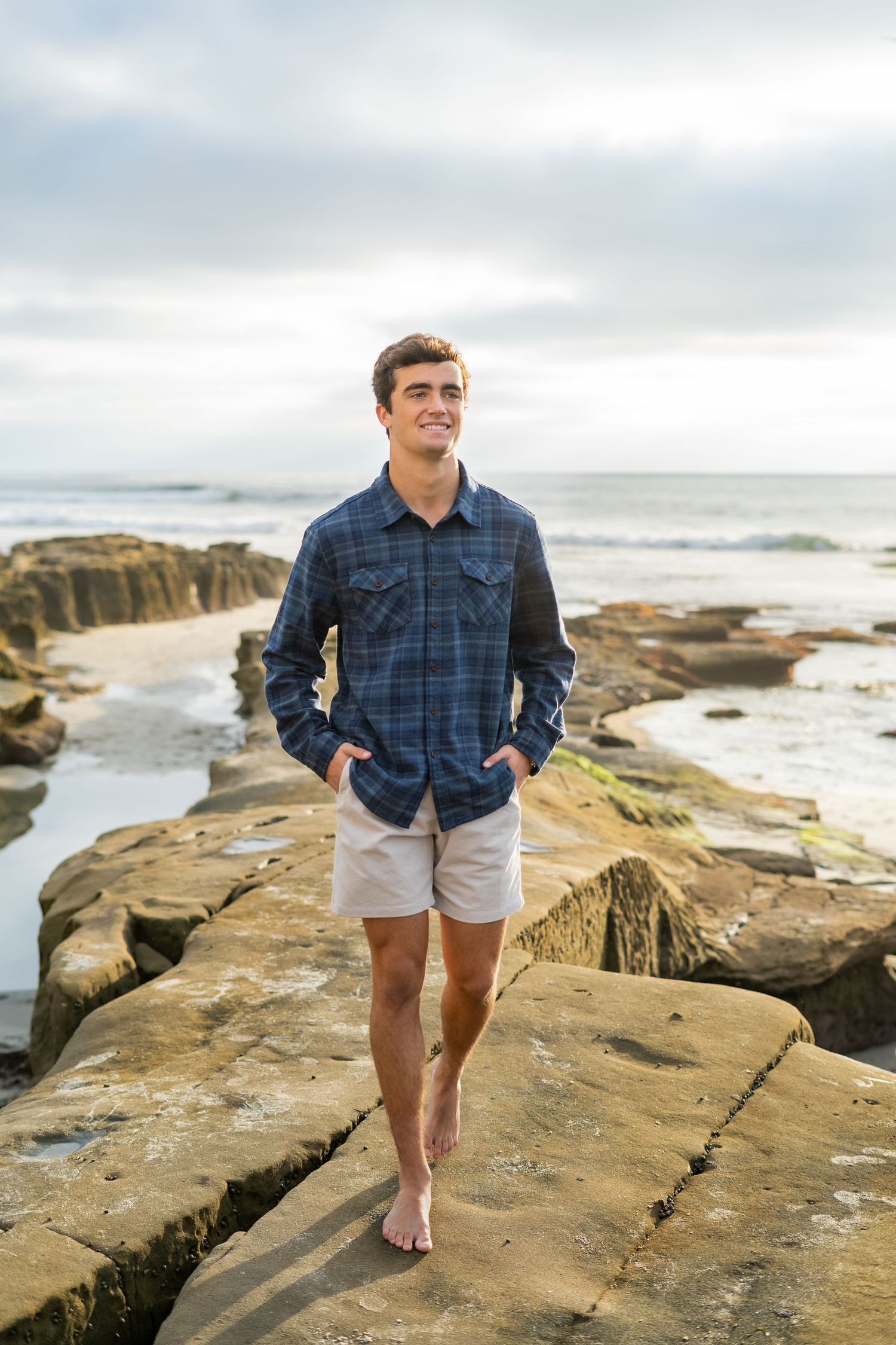 A high school senior in plaid shirt and shorts standing on rocky coastal cliffs at sunset.