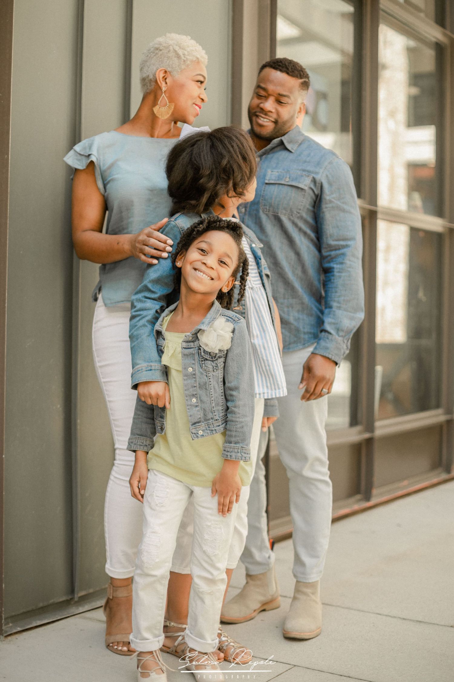 Playful family wearing coordinated denim and white outfits posing together against a building wall.