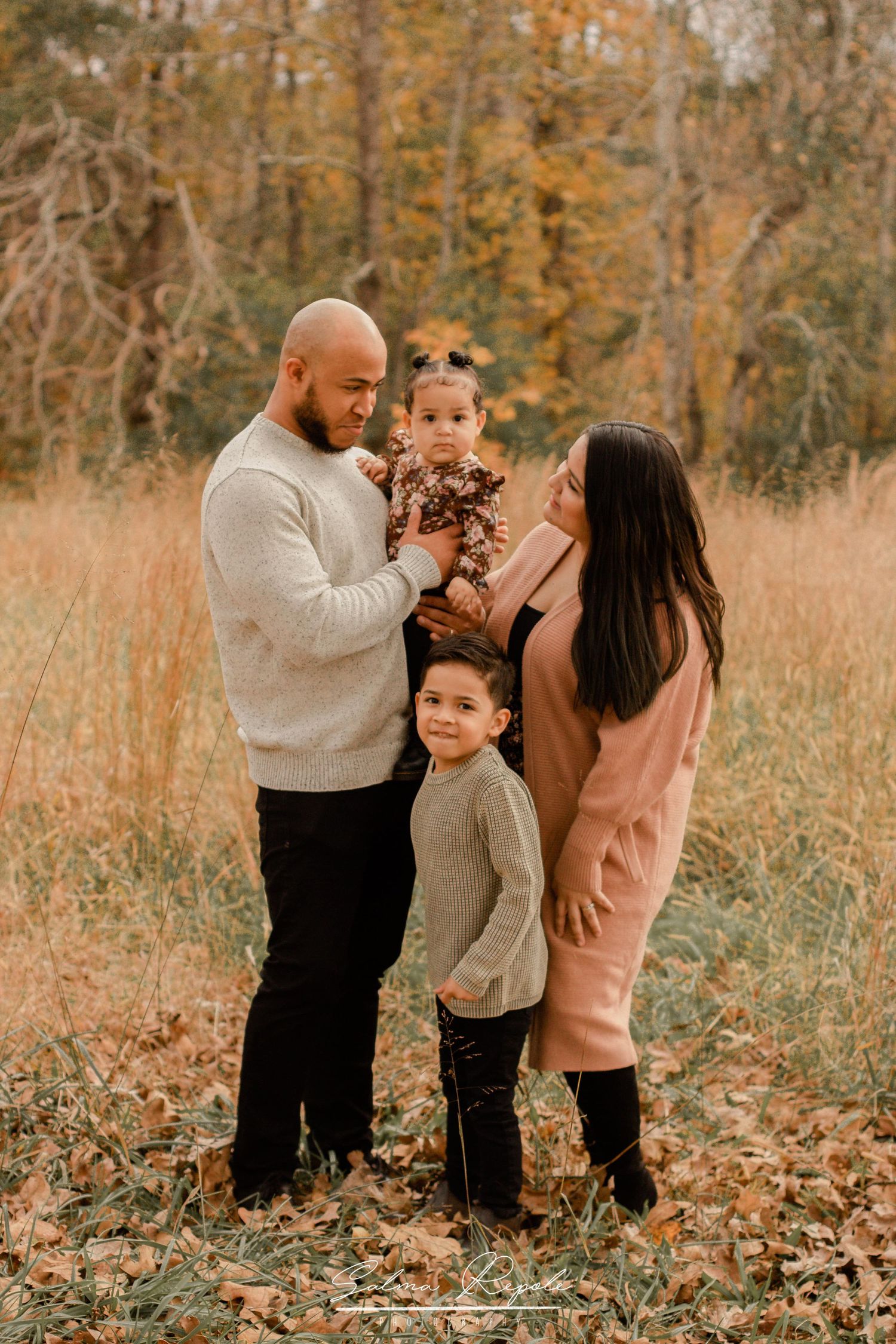 Family wearing coordinated outfits poses together in a golden autumn field setting.