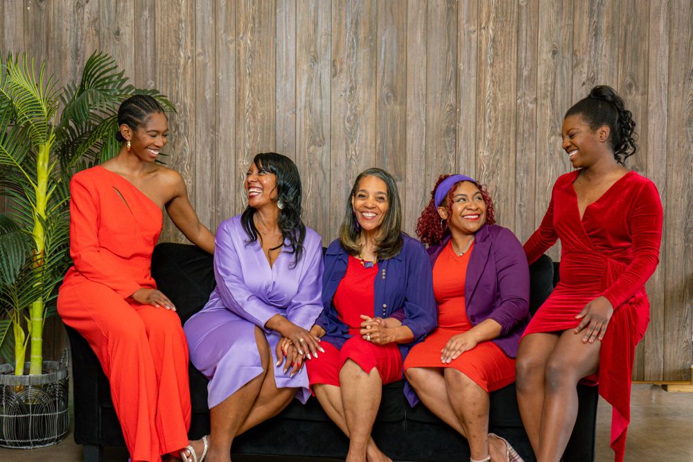 Five women in vibrant red and purple outfits laughing together on a stylish sofa.