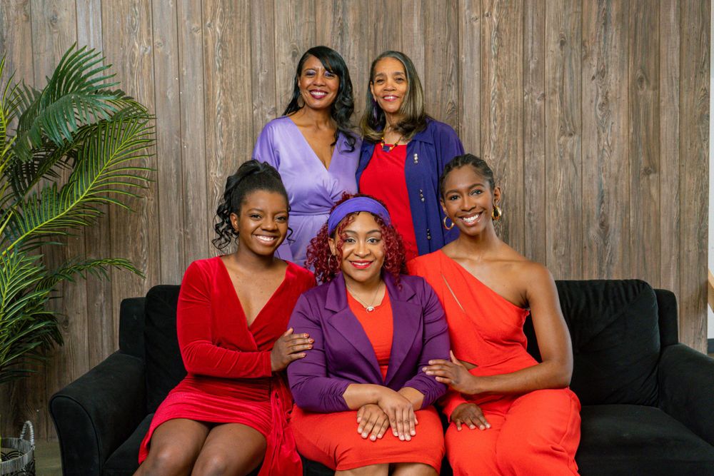 Five women in red and purple dresses posing for an elegant group portrait on a sofa.