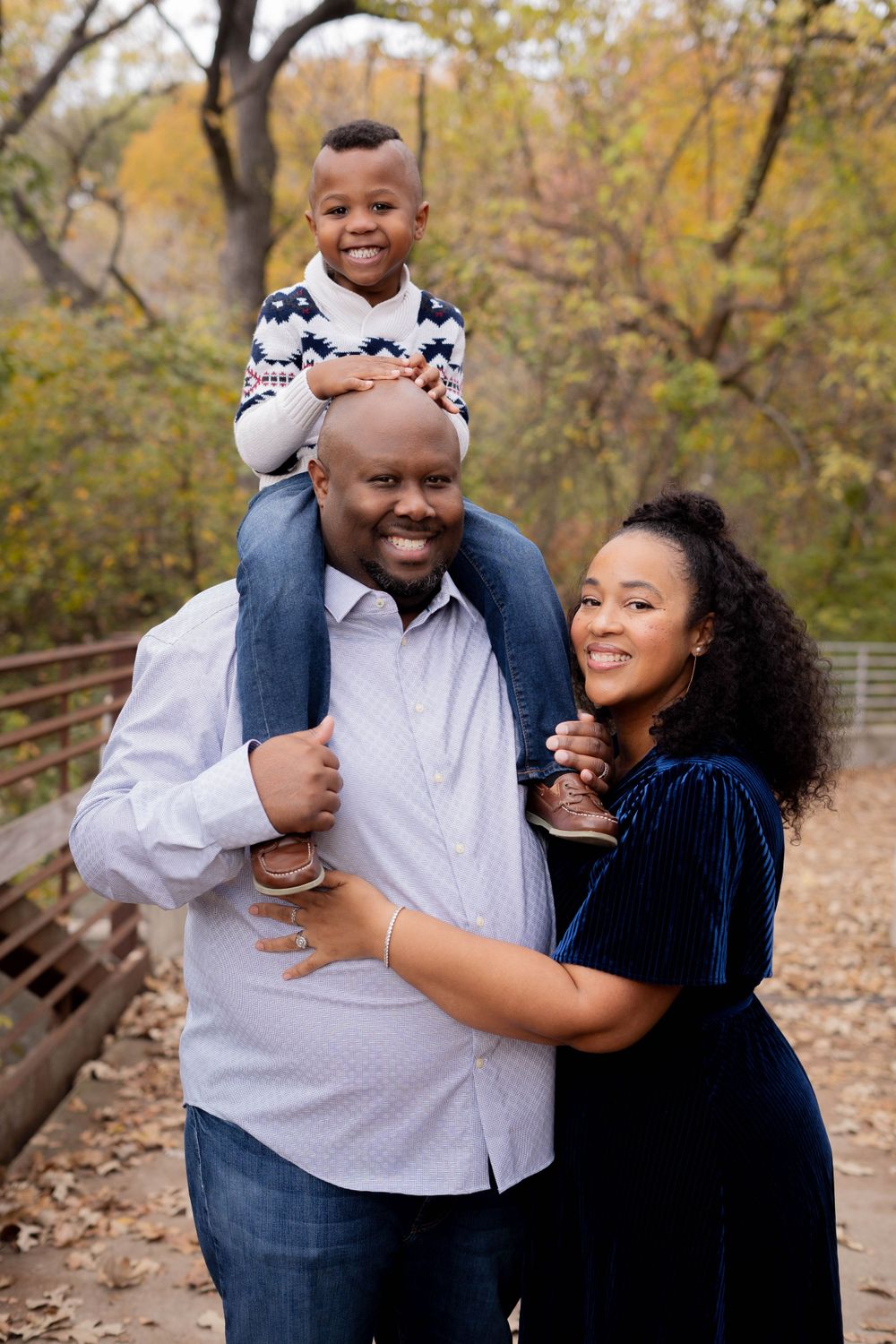 Father carries young son on shoulders while mother embraces them on a fall bridge.