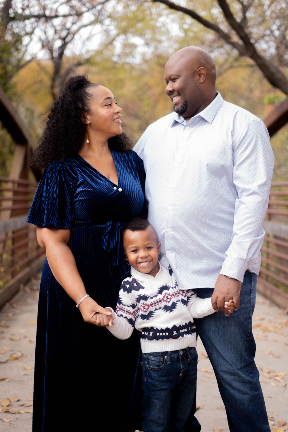 Smiling family of three holds hands on a bridge during an autumn outdoor photo session.