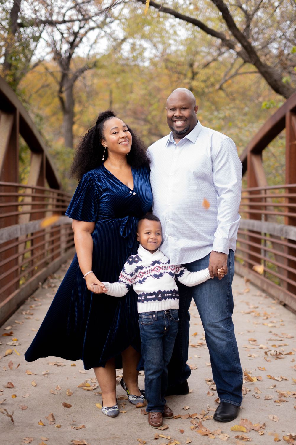 Happy family of three poses on a wooden bridge surrounded by autumn foliage.