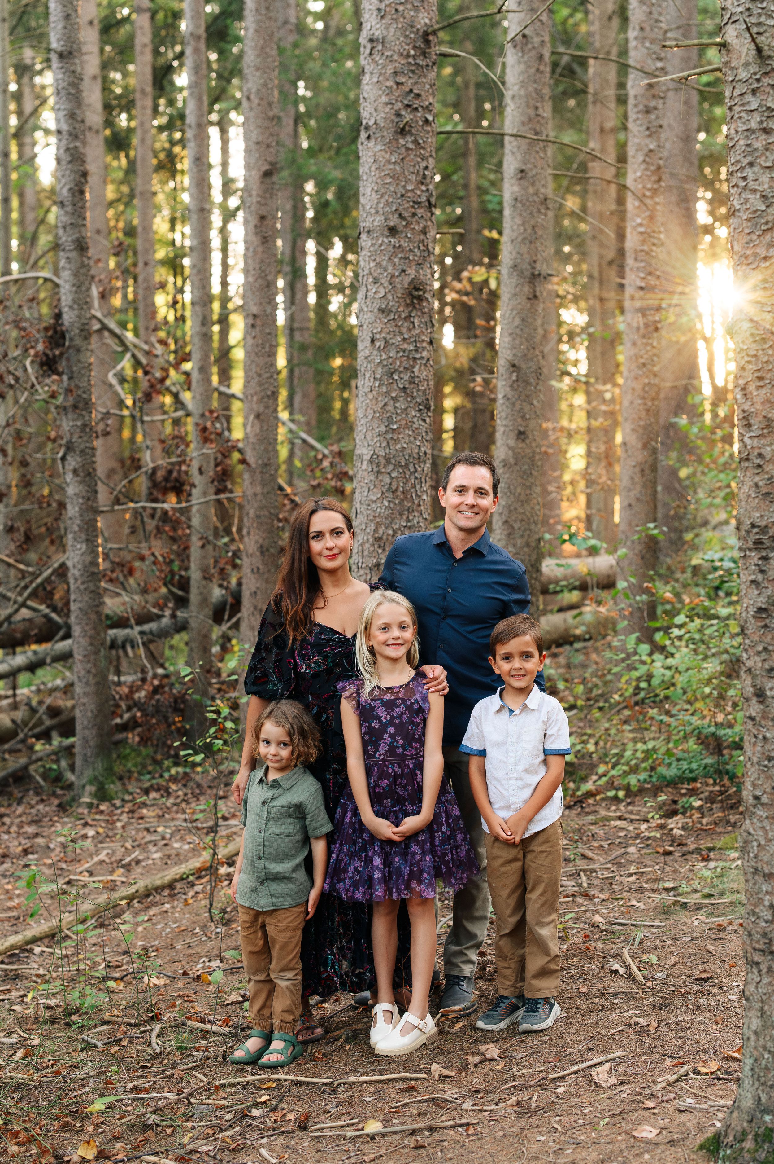 Family of five posing together in a sunlit forest, parents standing behind three young children on a woodland trail.