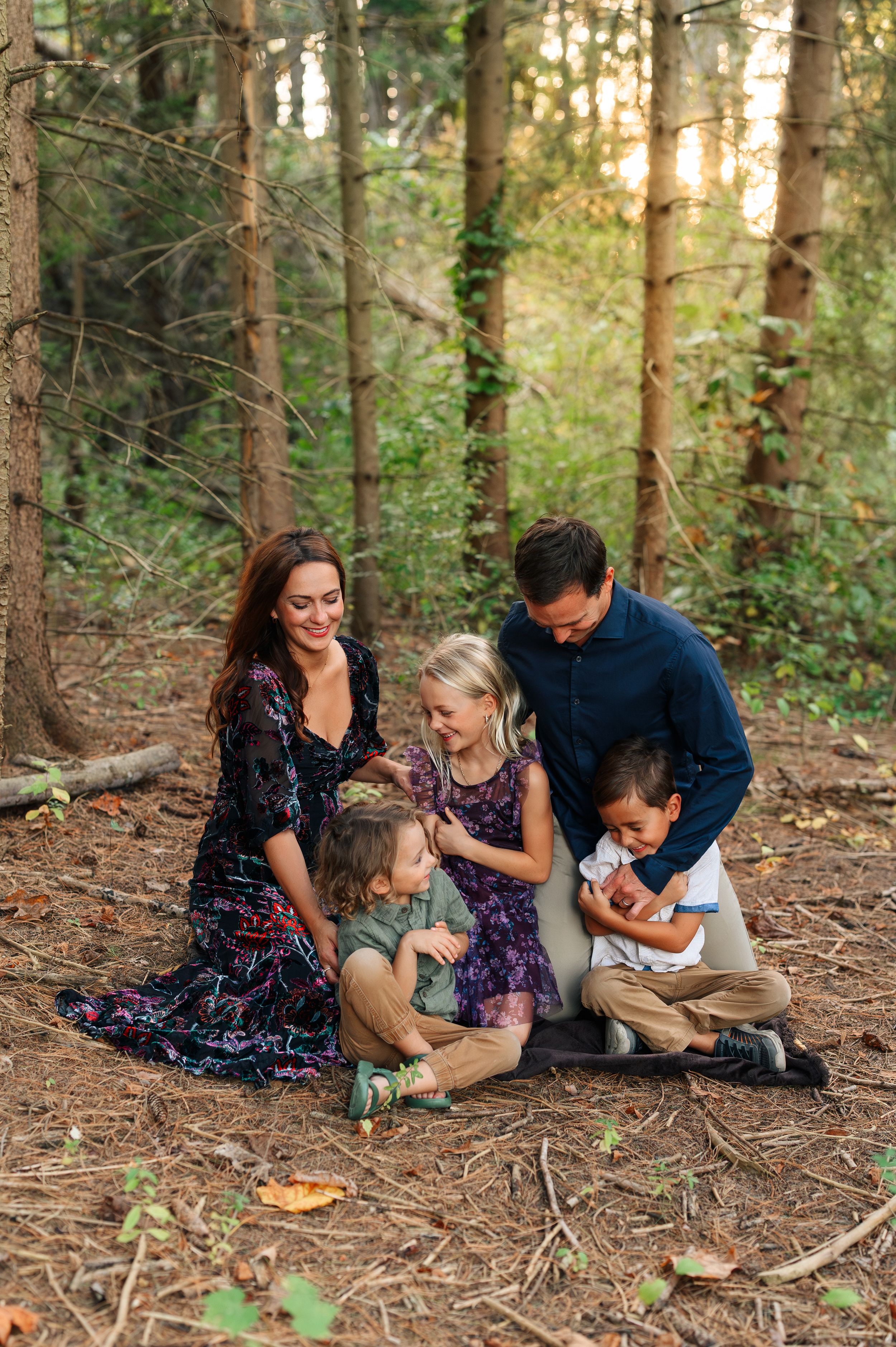 Family of five sharing a joyful moment together in a sunlit forest, sitting on pine needle-covered ground.
