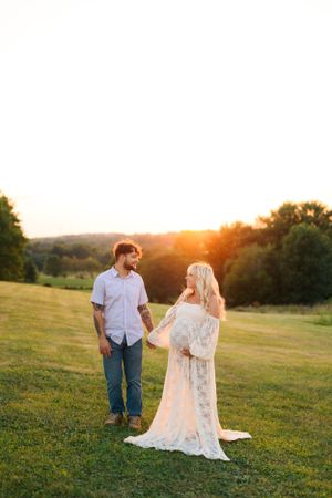 A couple stands in a sunlit field at sunset, with her wearing a flowing lace dress.