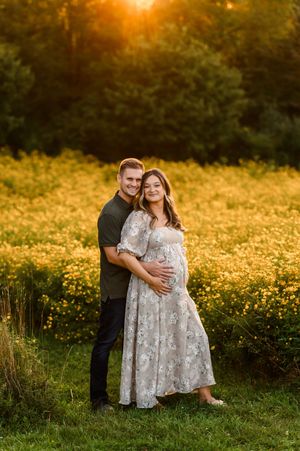 Couple poses in golden hour sunlight beside yellow wildflowers, woman in floral dress cradling baby bump during maternity photo session.