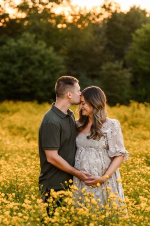 Couple sharing a tender moment in a golden field of yellow wildflowers during a maternity photo session at sunset.