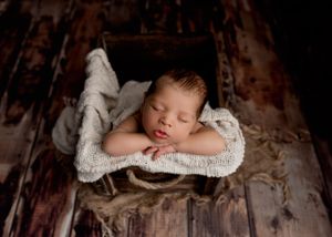 Sleeping newborn nestled in white knit blanket against rustic wooden backdrop.