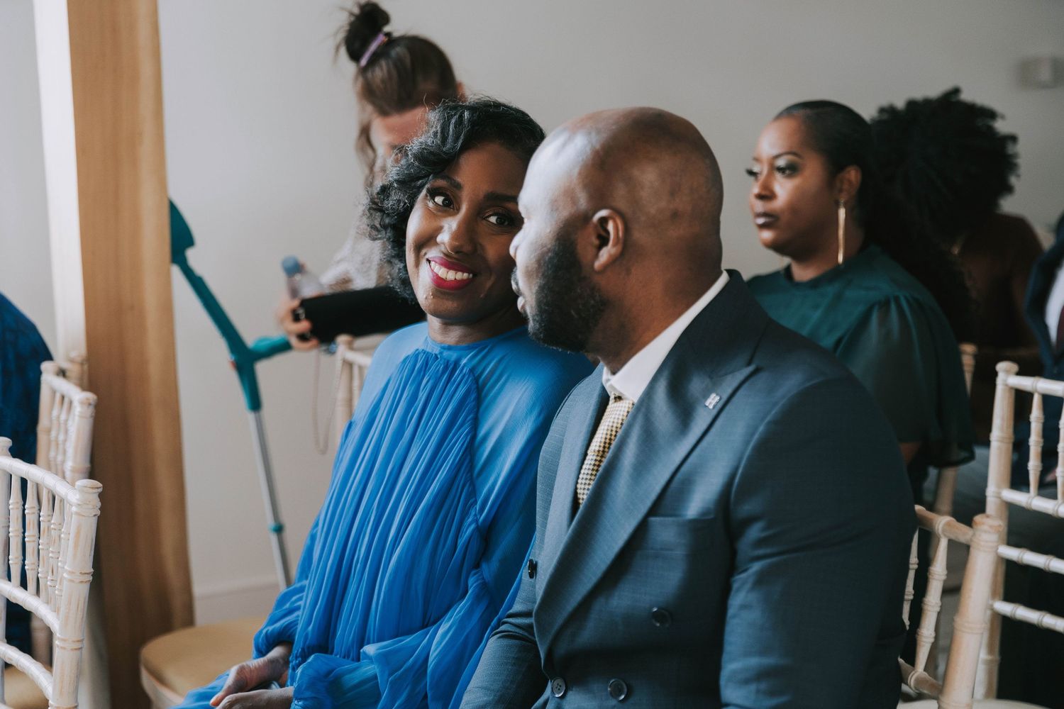 Wedding guests in coordinated blue attire seated in white chairs during an indoor ceremony.
