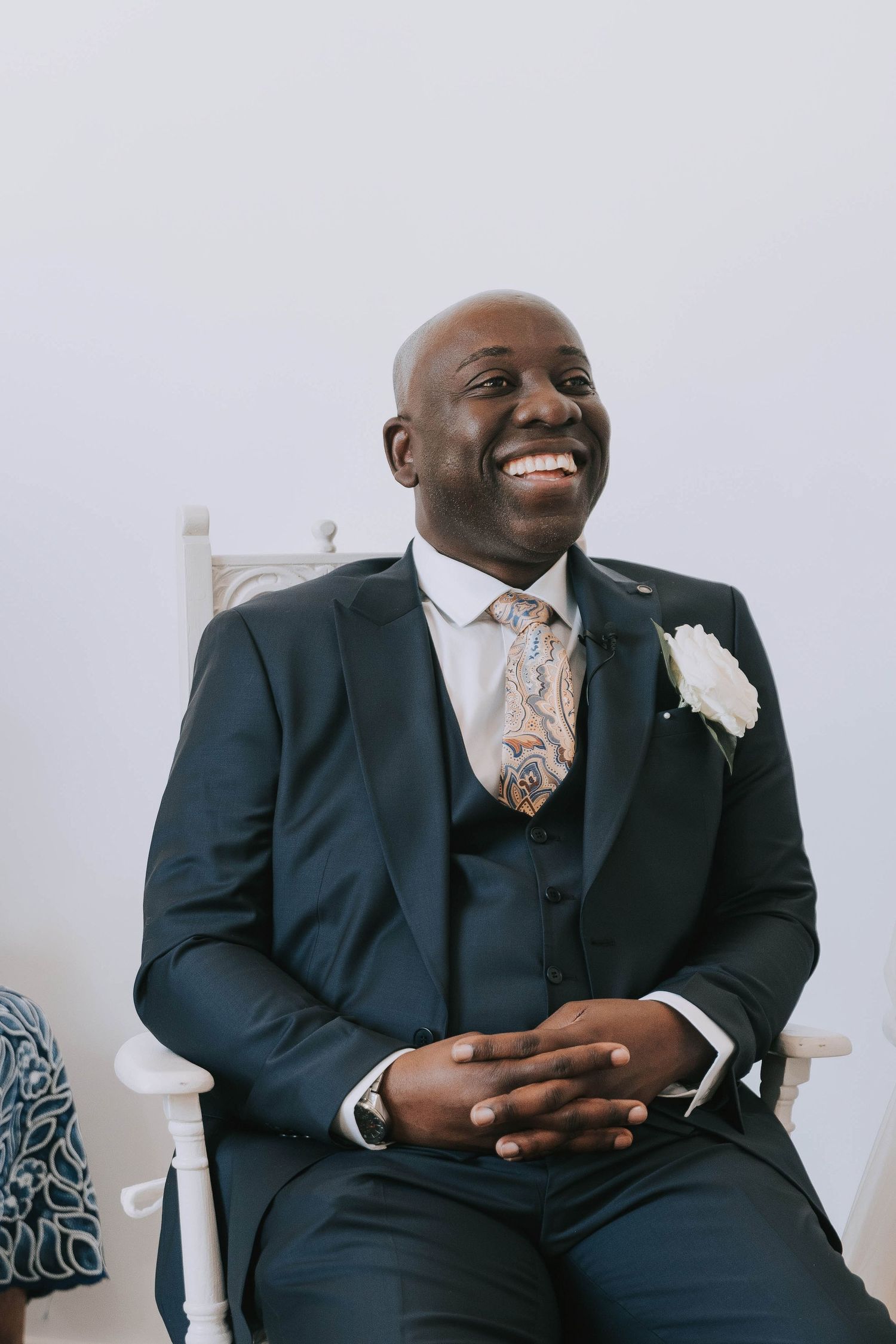 Groom in navy suit smiles while seated during wedding ceremony.