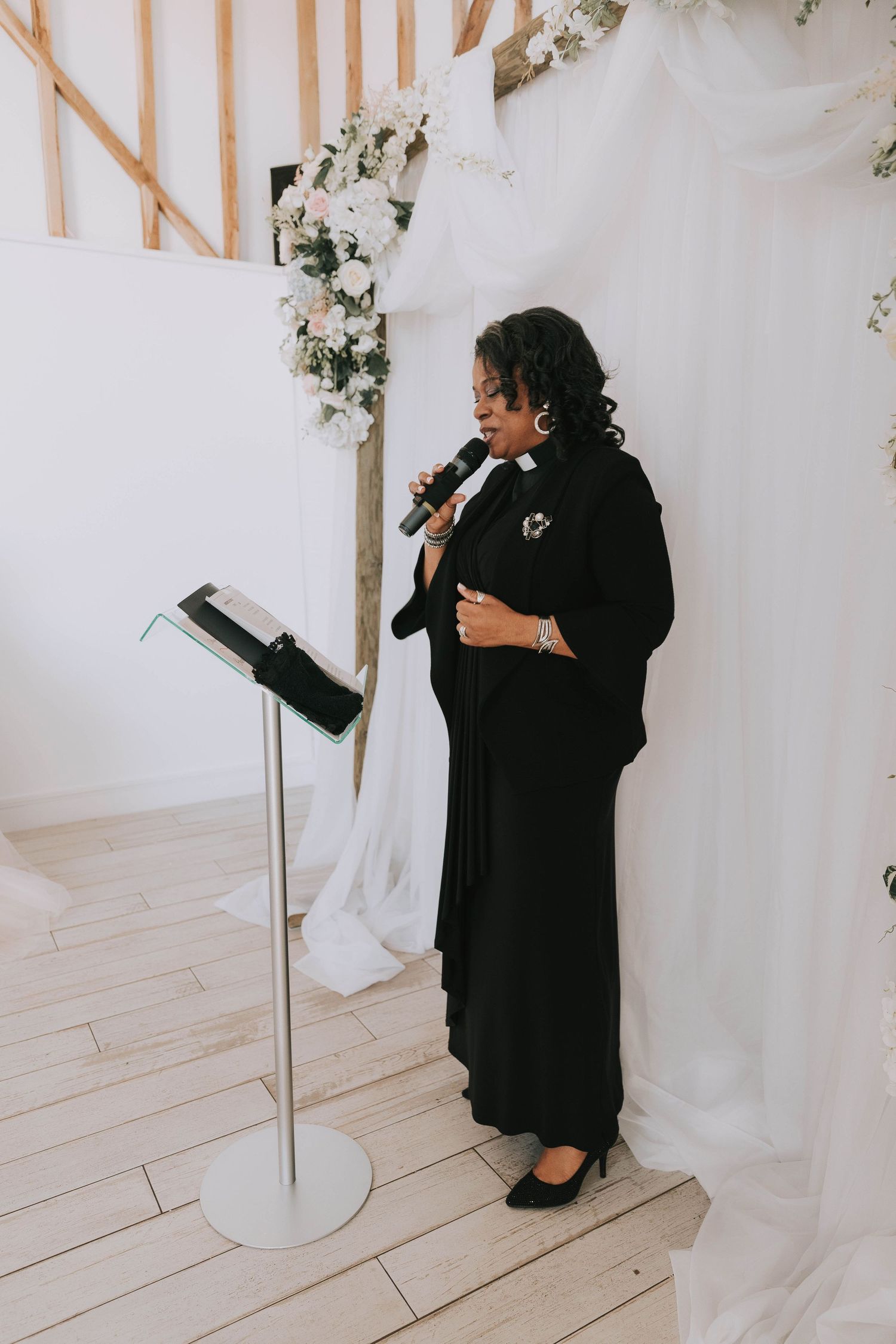 Wedding officiant in black holding a book during ceremony against white backdrop.