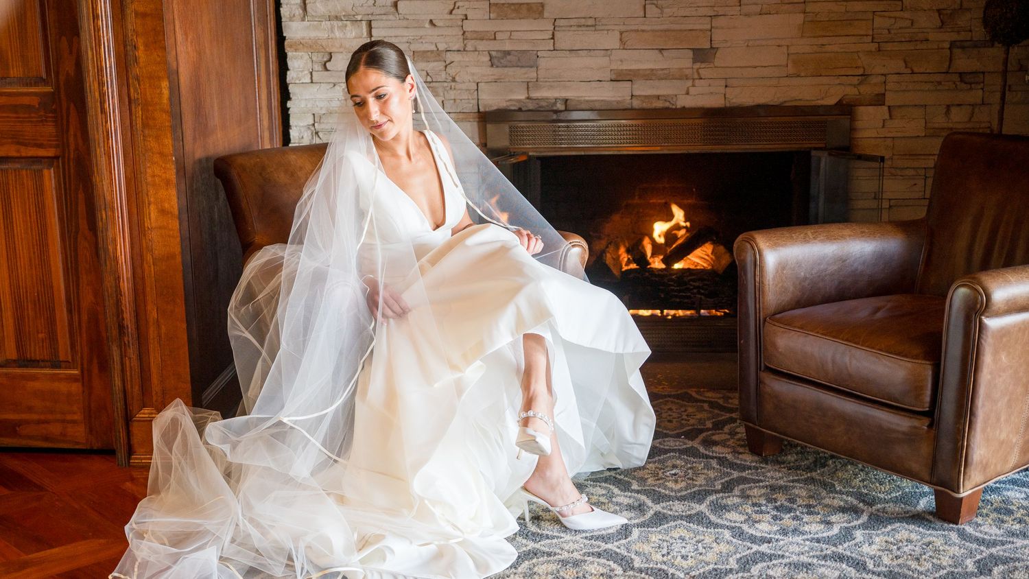 A person in an elegant white wedding gown and veil sits beside a cozy fireplace on a patterned area rug.