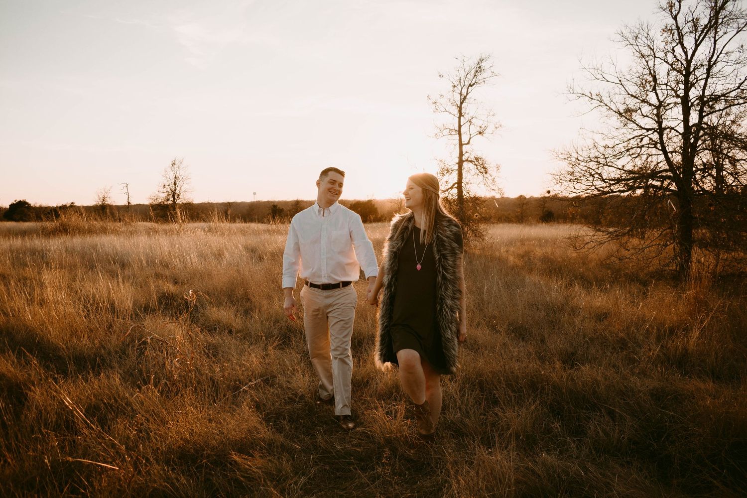 Couple walking together through tall grass during golden hour with bare trees in background.