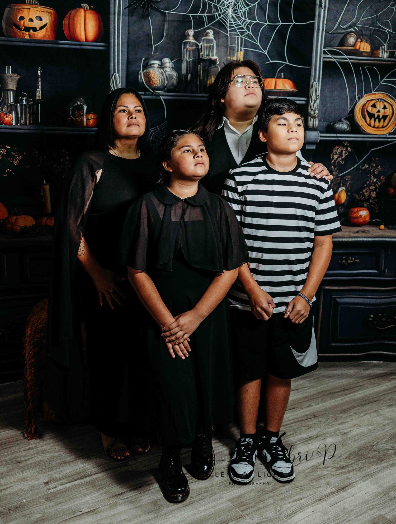 A group wearing dark colors poses in front of a Halloween themed backdrop with pumpkins and cobwebs.