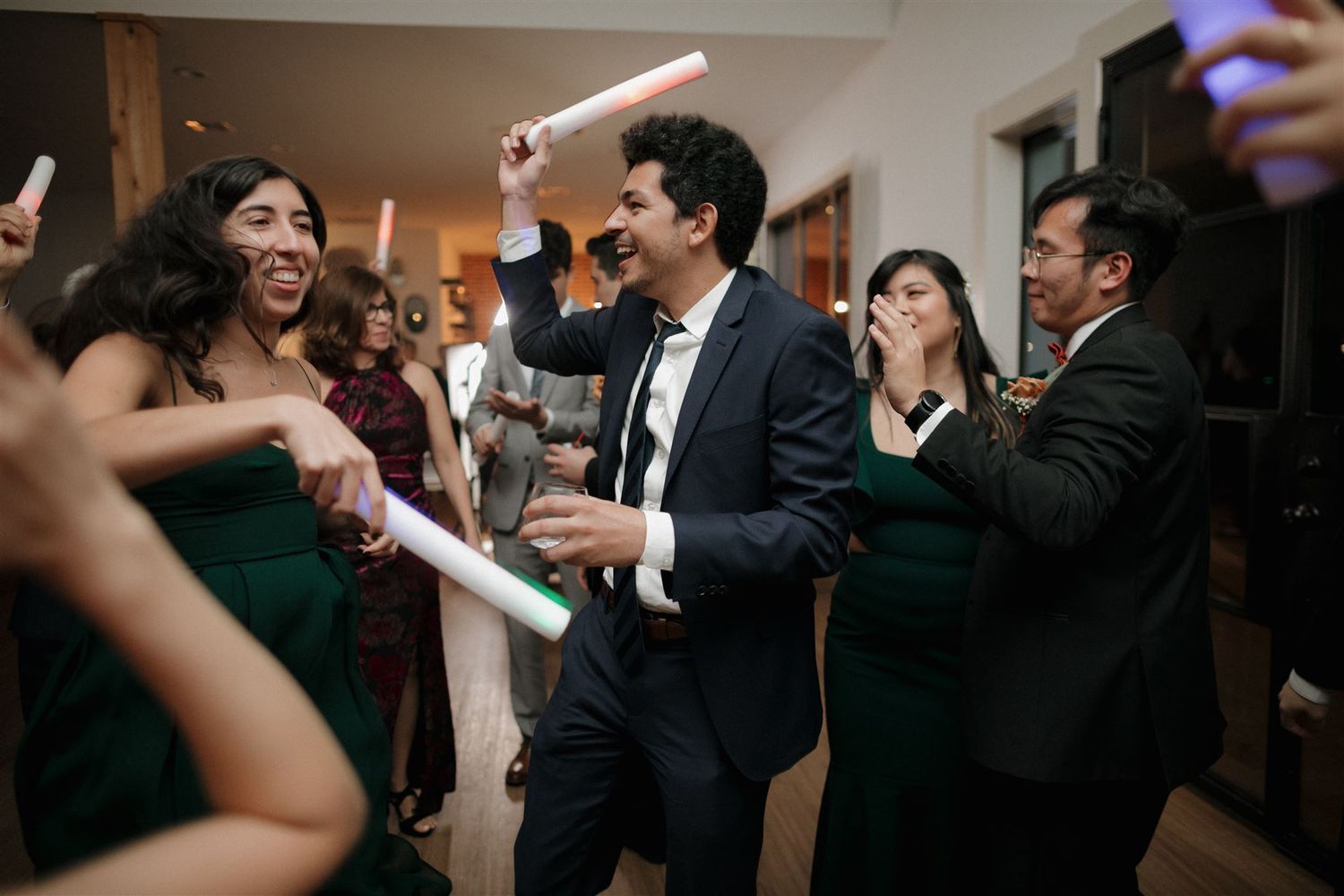 Party guests holding glow sticks while dancing and celebrating at an indoor event.