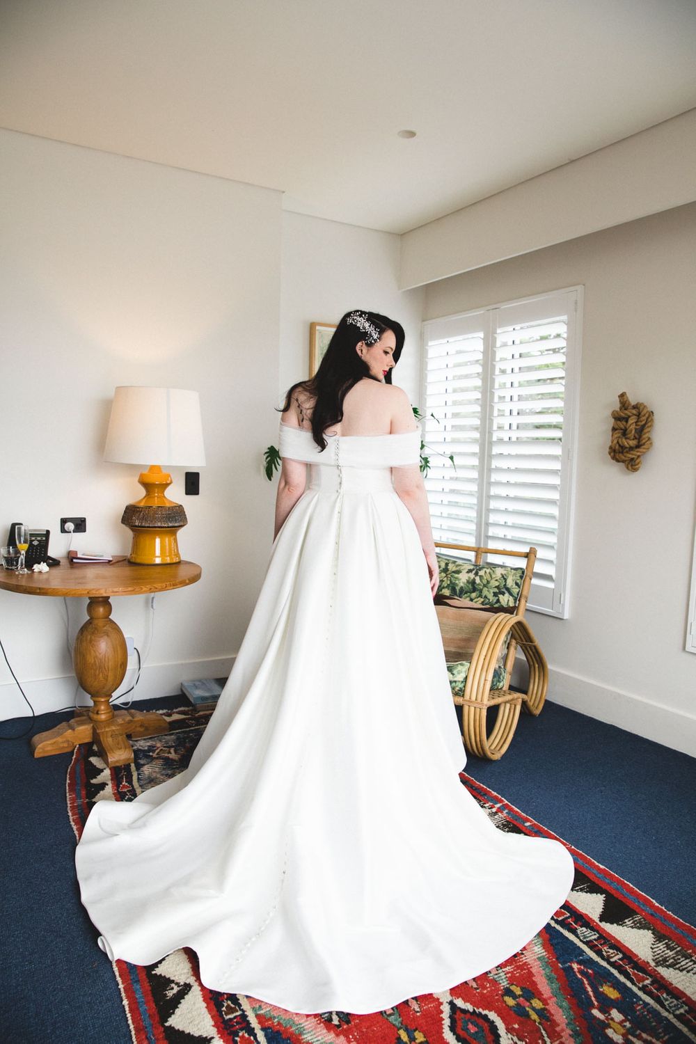 Bride in flowing white wedding dress photographed from behind in bright room with blue carpet.